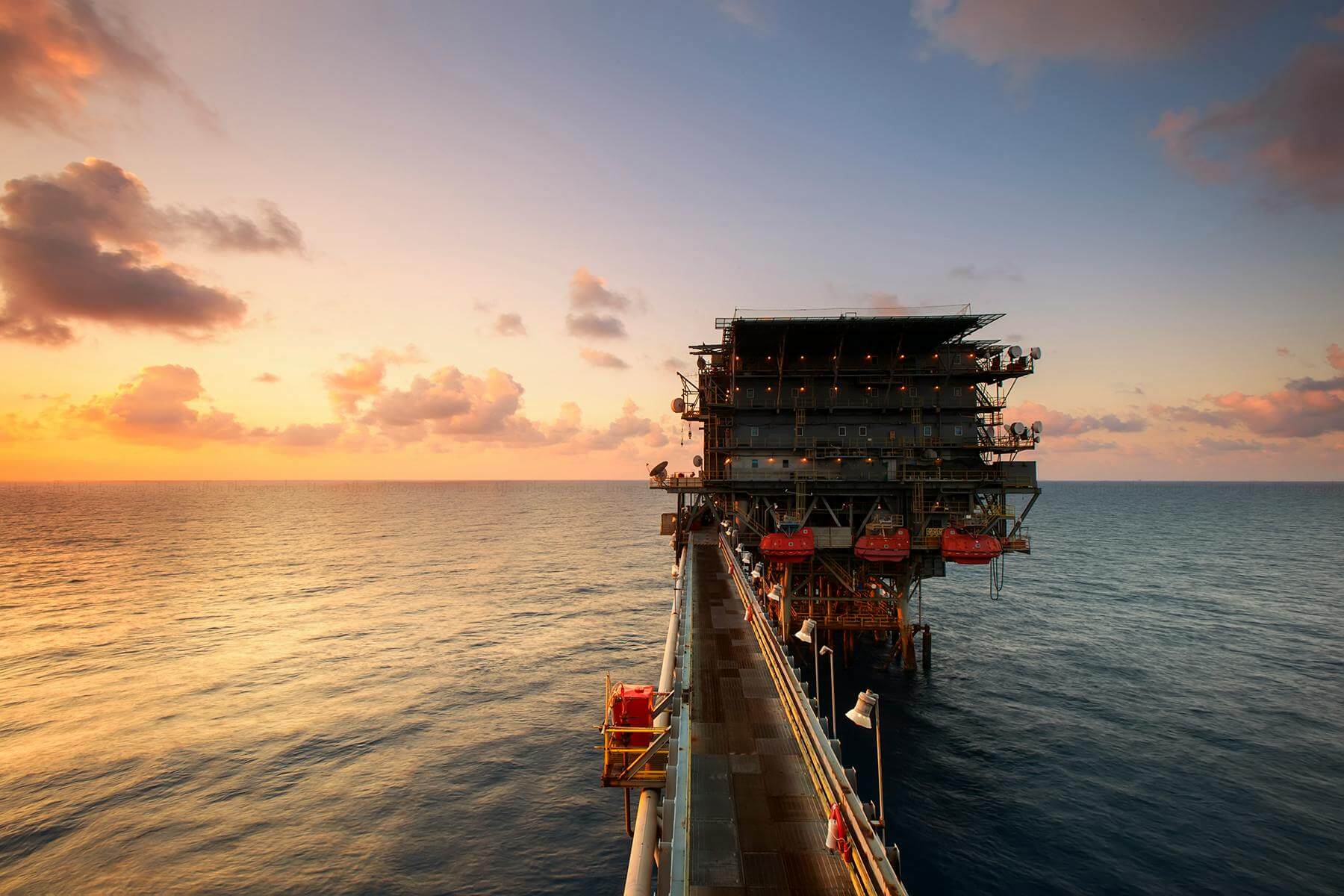 A serene sunset paints the sky with warm hues as the vast ocean surrounds an offshore oil rig, a testament to industrial might amidst the calm of nature.