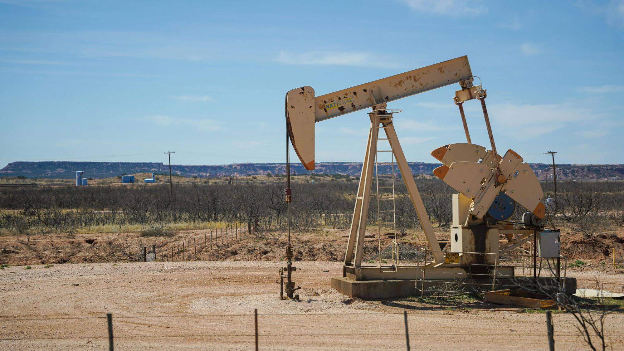 An oil pumpjack, also known as a nodding donkey, steadily operates in a barren field under a clear blue sky, emblematic of fossil fuel extraction in a rural landscape.