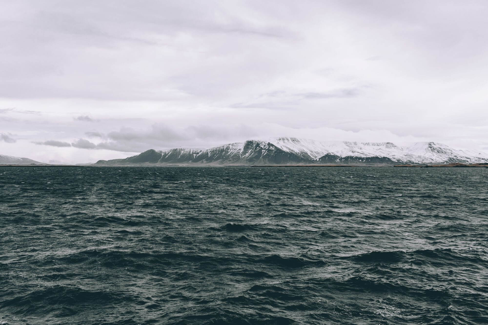 Choppy waters leading to a tranquil view of snow-capped mountains beneath a cloudy sky.