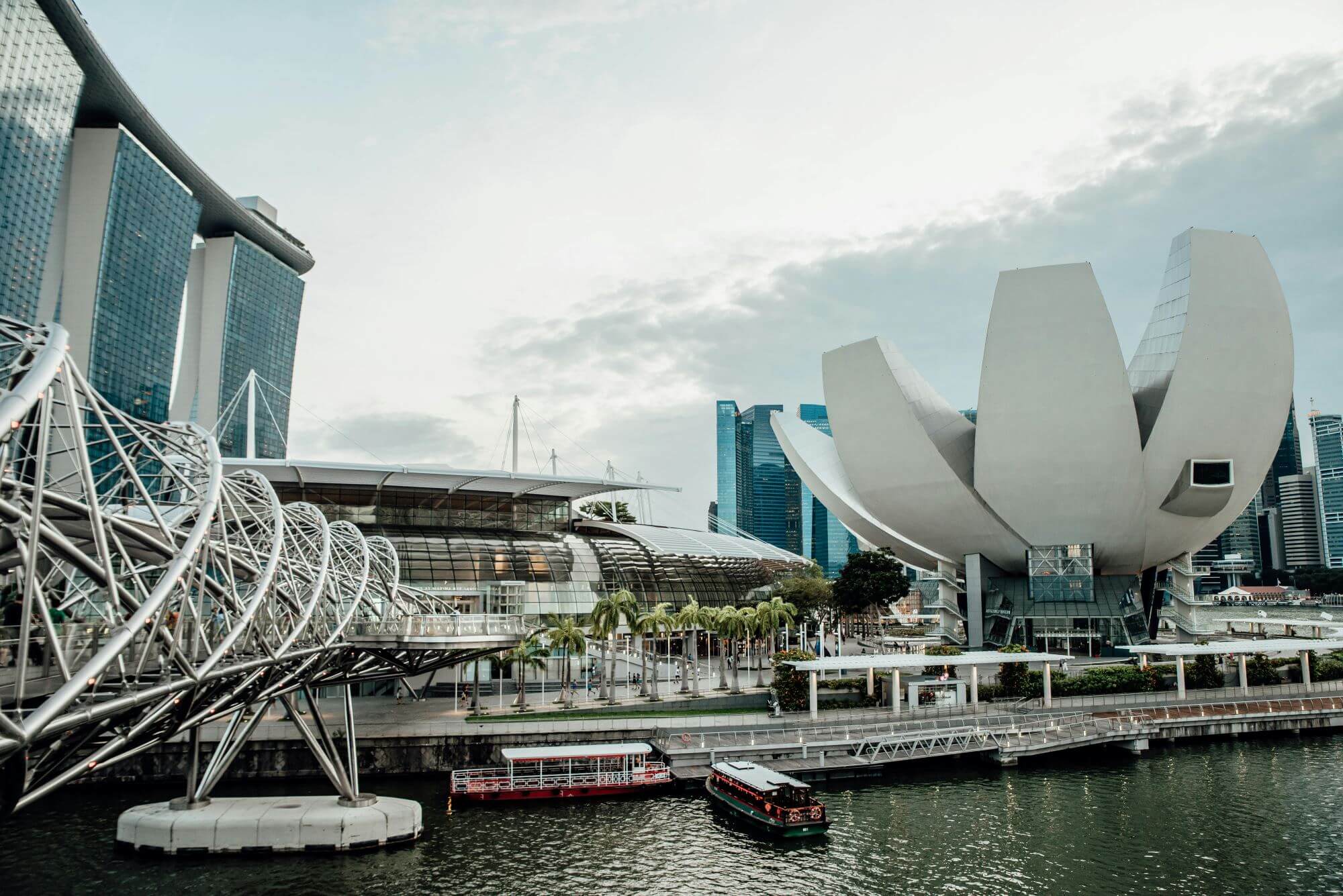 A view of the iconic marina bay in singapore featuring futuristic architecture including the artscience museum resembling a lotus flower, the distinctive helix bridge, and towering skyscrapers against a cloudy sky. a tour boat cruises along the serene waters of the bay.