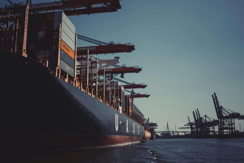 Cargo ship loaded with containers docked at a busy industrial port with cranes ready for unloading during dusk.