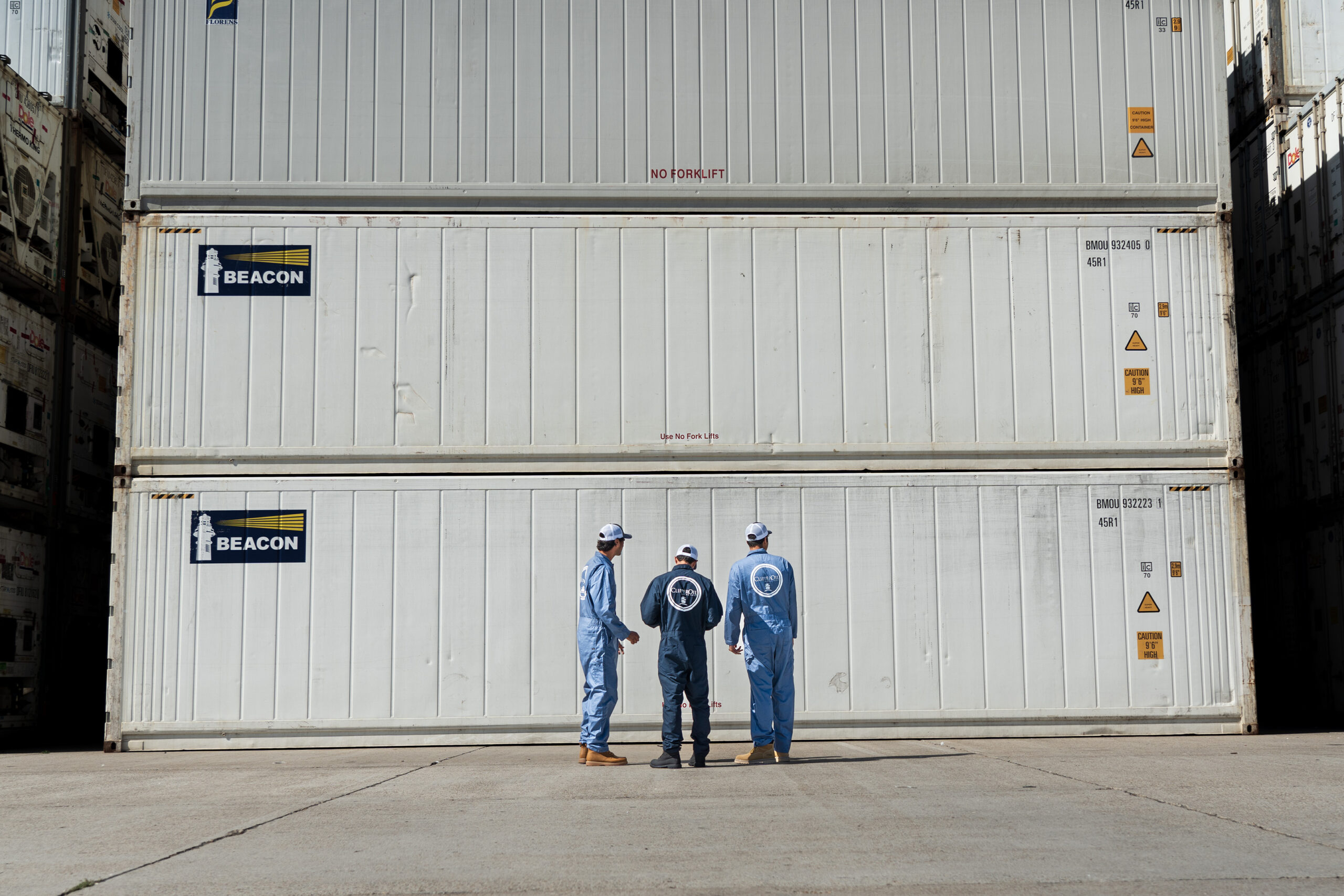 Three workers in safety gear standing in front of large cargo containers at a shipping yard discussing operations related to marine bunker fuel prices.