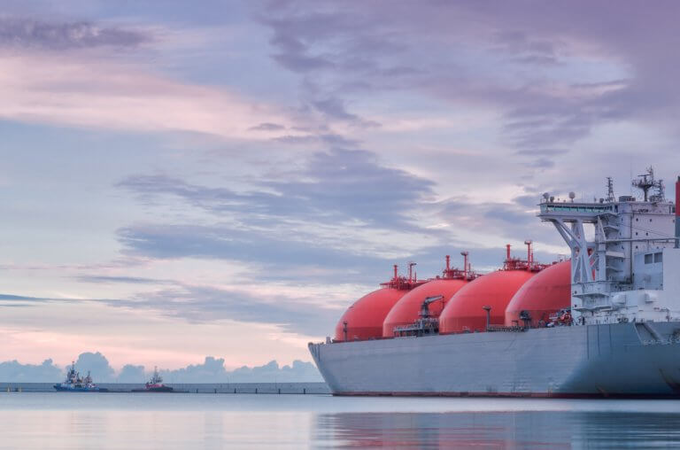 A large LNG tanker with round red storage tanks is docked at sea under a cloudy, pastel-colored sky. Smaller boats are visible in the distance near the horizon.