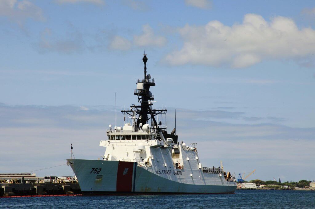 A U.S. Coast Guard ship, numbered 752, is docked at a pier on a sunny day. The vessel is white with red and blue stripes and has antennas and radar equipment on its mast. The sky is mostly clear with some clouds.