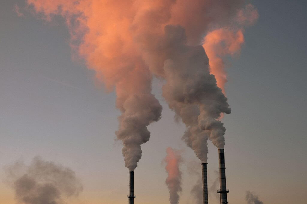 Four industrial smokestacks release thick plumes of smoke into the sky at sunset, with the clouds tinged orange and pink by the fading sunlight.