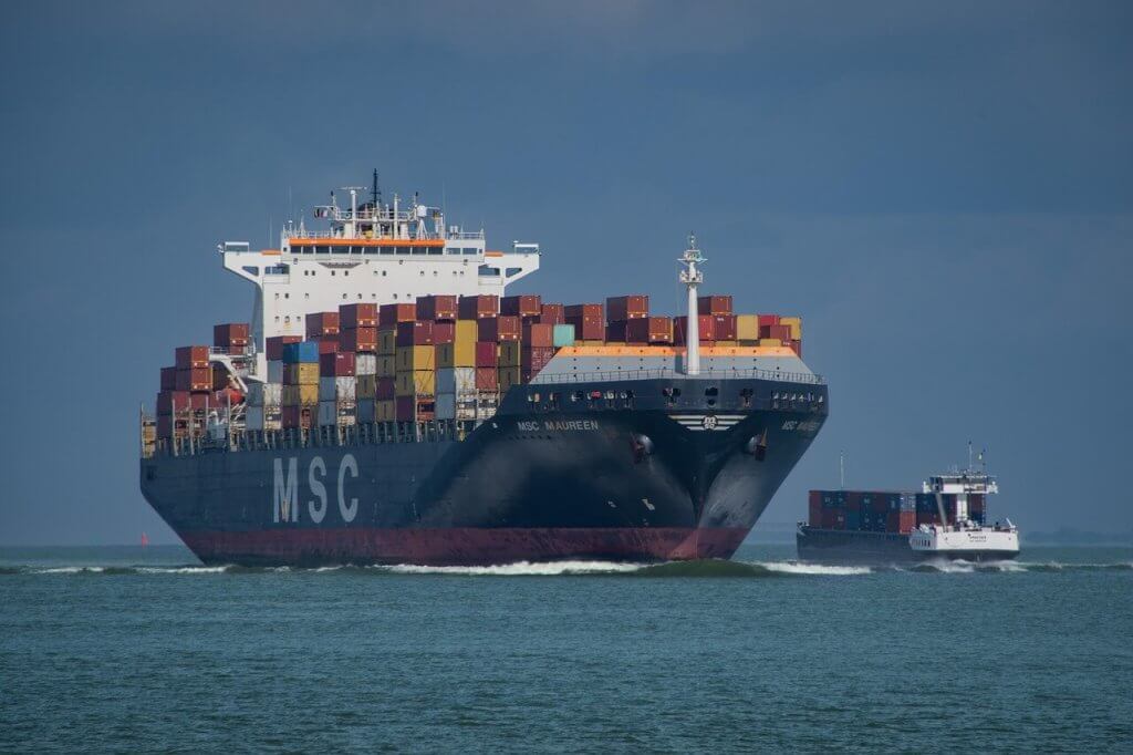 A large MSC container ship loaded with colorful shipping containers sails on the ocean, accompanied by a smaller cargo vessel to its right under a cloudy sky.