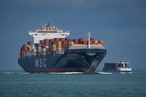 A large MSC container ship loaded with colorful shipping containers sails on the ocean, accompanied by a smaller cargo vessel to its right under a cloudy sky.