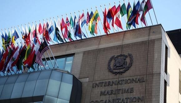 A building with the inscription "International Maritime Organization" on its wall, displaying many international flags on poles along the roofline against a clear blue sky.
