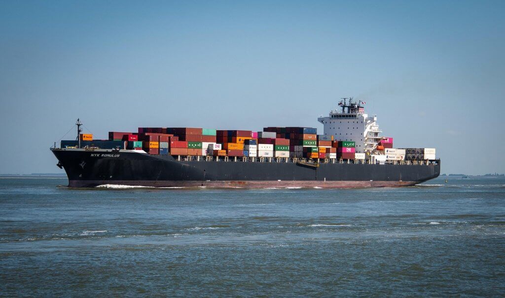 A large cargo ship loaded with colorful shipping containers sails on calm water under a clear blue sky.
