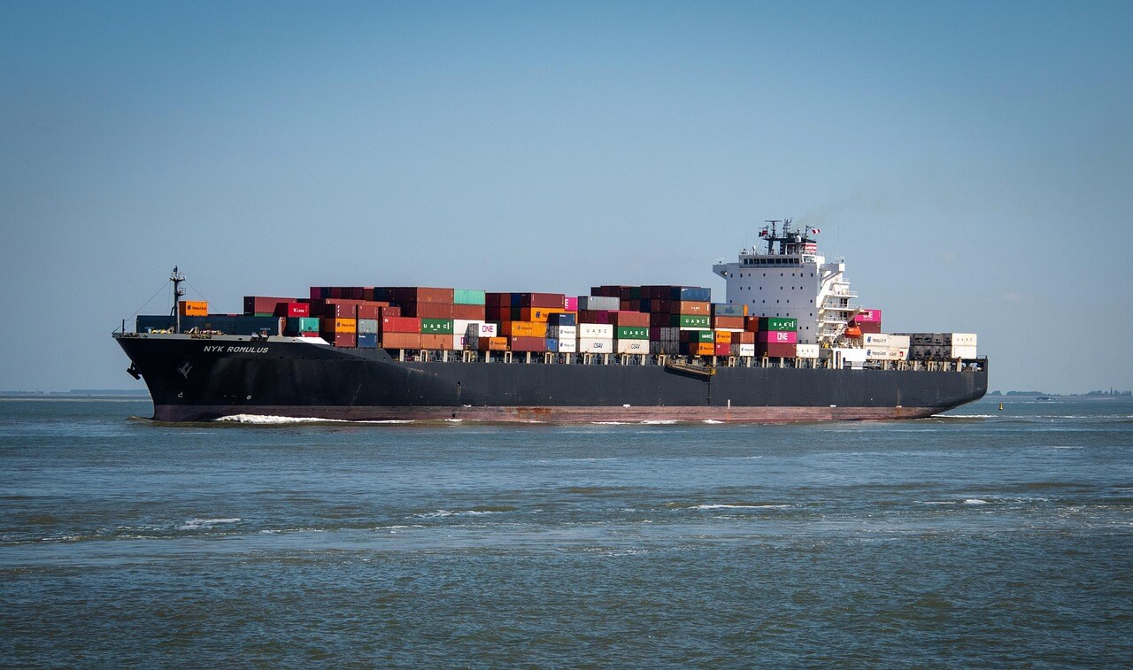 A large cargo ship loaded with colorful shipping containers sails on calm water under a clear blue sky.