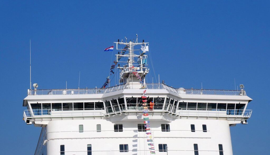 The image shows the control bridge of a large white ship decorated with colorful signal flags, set against a clear blue sky. The ship appears modern and is viewed from the front, highlighting its upper deck and windows.
