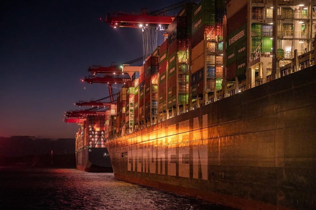 Two large container ships, brightly lit and loaded with colorful shipping containers, are docked side by side at a port at night. Cranes extend over the ships, and the water reflects the lights.