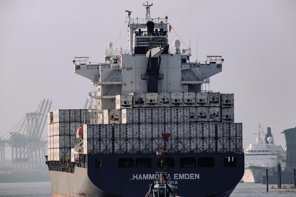 A large cargo ship loaded with stacked shipping containers sails in a harbor, with cranes and another ship visible in the background under a hazy sky.