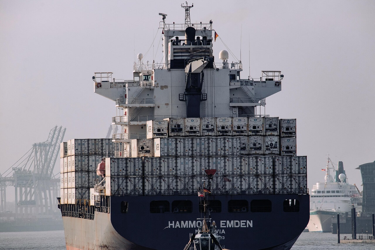 A large cargo ship loaded with stacked shipping containers sails in a harbor, with cranes and another ship visible in the background under a hazy sky.
