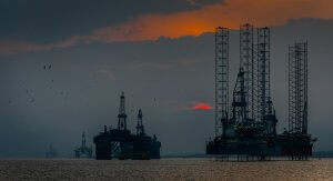 Offshore oil rigs silhouetted against a dramatic sunset sky, with dark clouds and the sun partially visible near the horizon. Birds fly over the calm sea in the foreground.