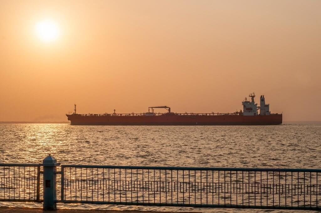 A large cargo ship sails on a calm sea during sunset, with the sun low in the sky and warm orange hues. A metal railing and concrete walkway are visible in the foreground.
