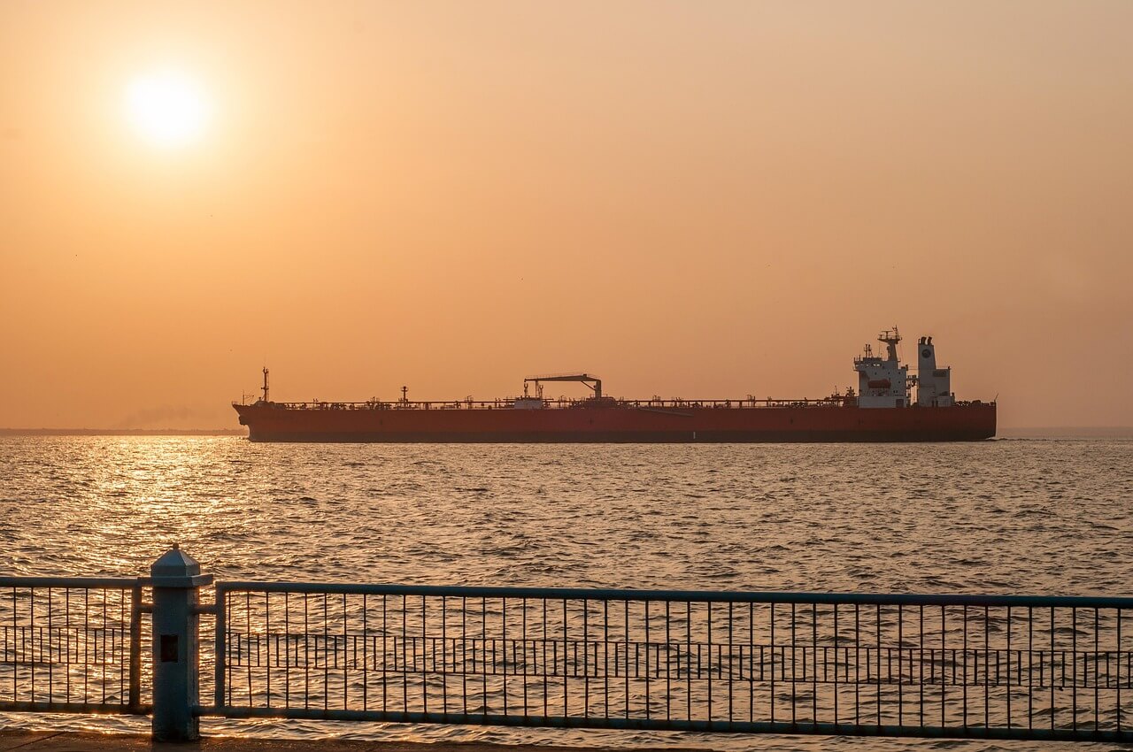 A large cargo ship sails on a calm sea during sunset, with the sun low in the sky and warm orange hues. A metal railing and concrete walkway are visible in the foreground.