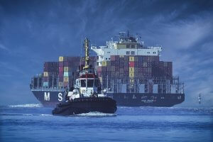 A large container ship loaded with colorful cargo containers sails in the ocean, followed closely by a smaller tugboat in the foreground under a cloudy blue sky.