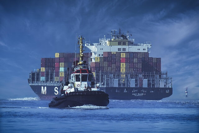 A large container ship loaded with colorful cargo containers sails in the ocean, followed closely by a smaller tugboat in the foreground under a cloudy blue sky.