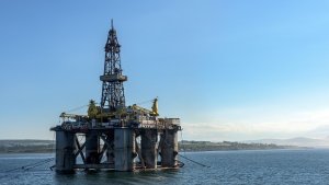 An offshore oil drilling platform stands in calm water near the coastline under a clear blue sky, with distant land and low hills visible in the background.