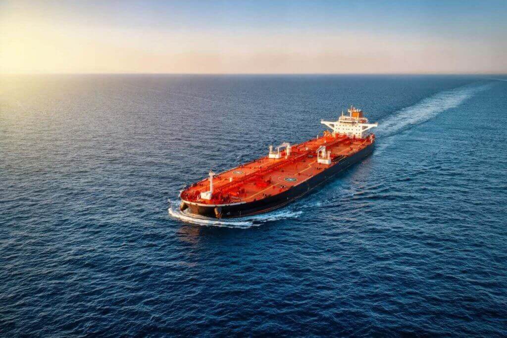 A large red oil tanker sails through calm blue ocean water under a clear sky, leaving a white wake behind it as the sun sets on the horizon.