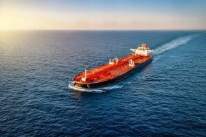 A large red oil tanker sails through calm blue ocean water under a clear sky, leaving a white wake behind it as the sun sets on the horizon.