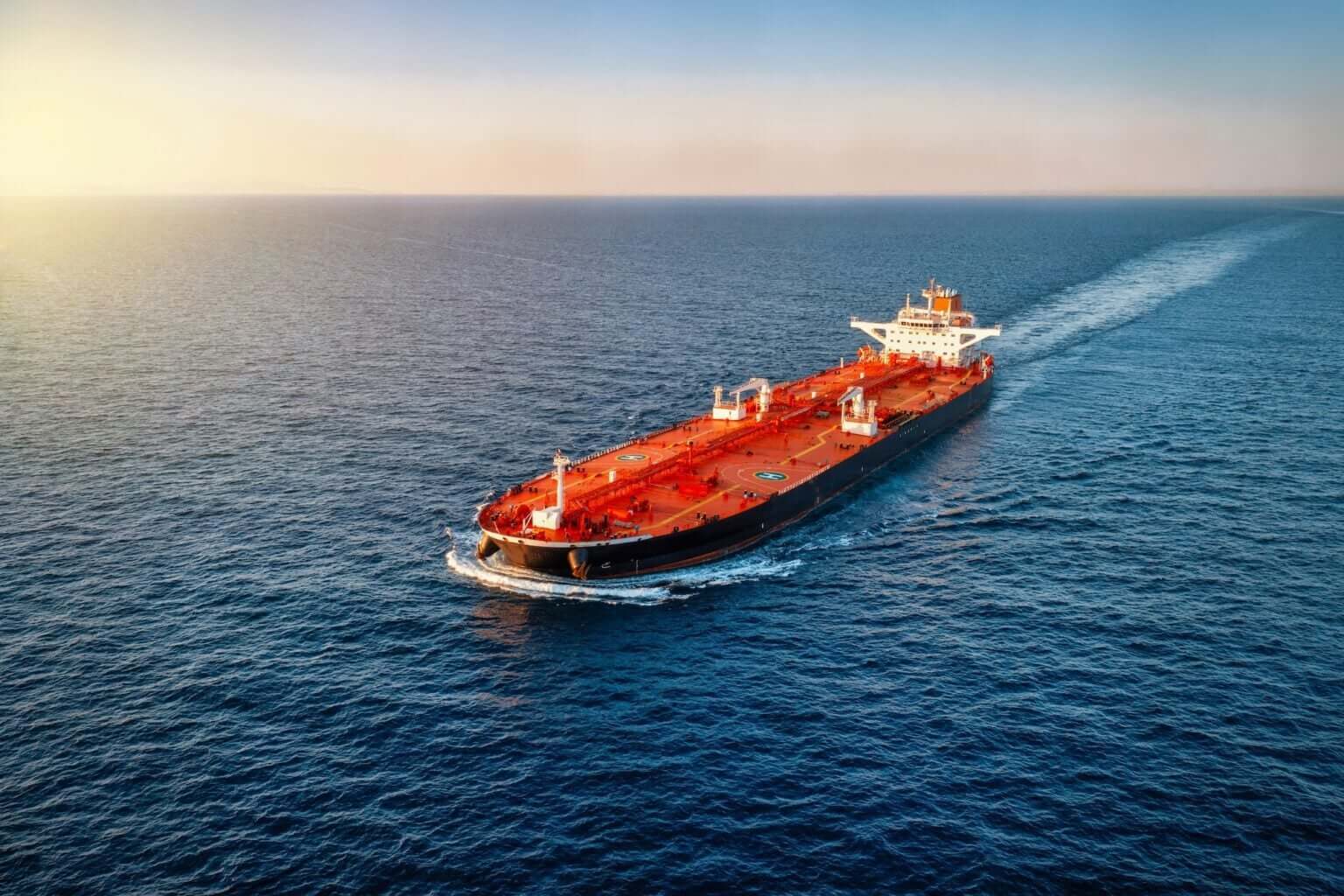 A large red oil tanker sails through calm blue ocean water under a clear sky, leaving a white wake behind it as the sun sets on the horizon.
