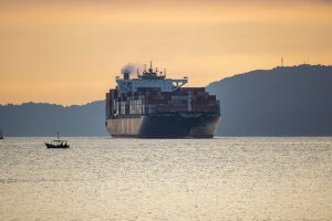 A large cargo ship with stacked containers sails on calm water at sunset, with a small boat nearby and a silhouette of hills in the background.