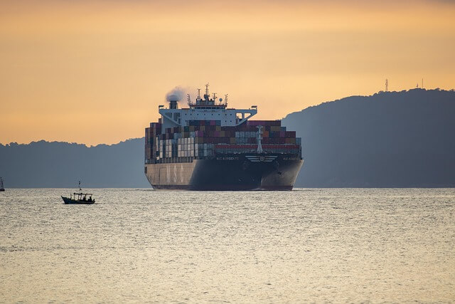 A large cargo ship with stacked containers sails on calm water at sunset, with a small boat nearby and a silhouette of hills in the background.