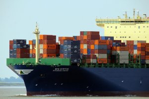 A large cargo ship loaded with colorful shipping containers sails on the water. The ship’s hull is dark blue and the name “HYUNDAI DRIVE” is visible. The sky is clear and calm.