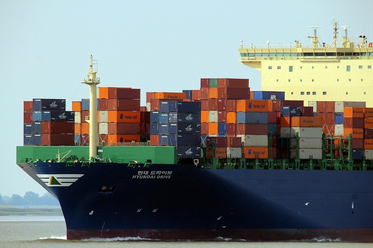 A large cargo ship loaded with colorful shipping containers sails on the water. The ship’s hull is dark blue and the name “HYUNDAI DRIVE” is visible. The sky is clear and calm.