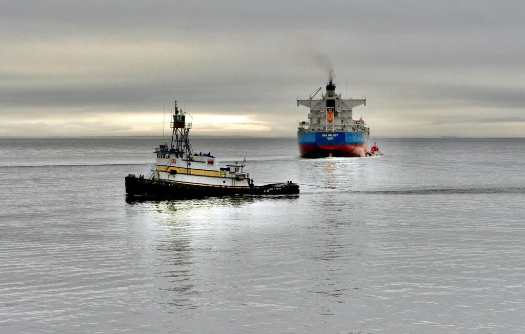 A small tugboat pulls a large cargo ship across a calm, gray sea under a cloudy sky, with the horizon faintly visible in the distance.