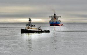 A small tugboat pulls a large cargo ship across a calm, gray sea under a cloudy sky, with the horizon faintly visible in the distance.