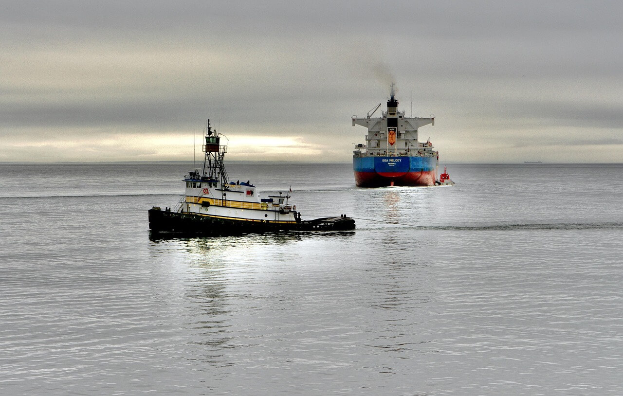 A small tugboat pulls a large cargo ship across a calm, gray sea under a cloudy sky, with the horizon faintly visible in the distance.