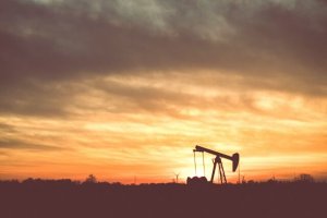 Oil pump jack silhouette in a field at sunset, with an orange and yellow sky and scattered clouds. Trees and wind turbines are visible on the horizon.