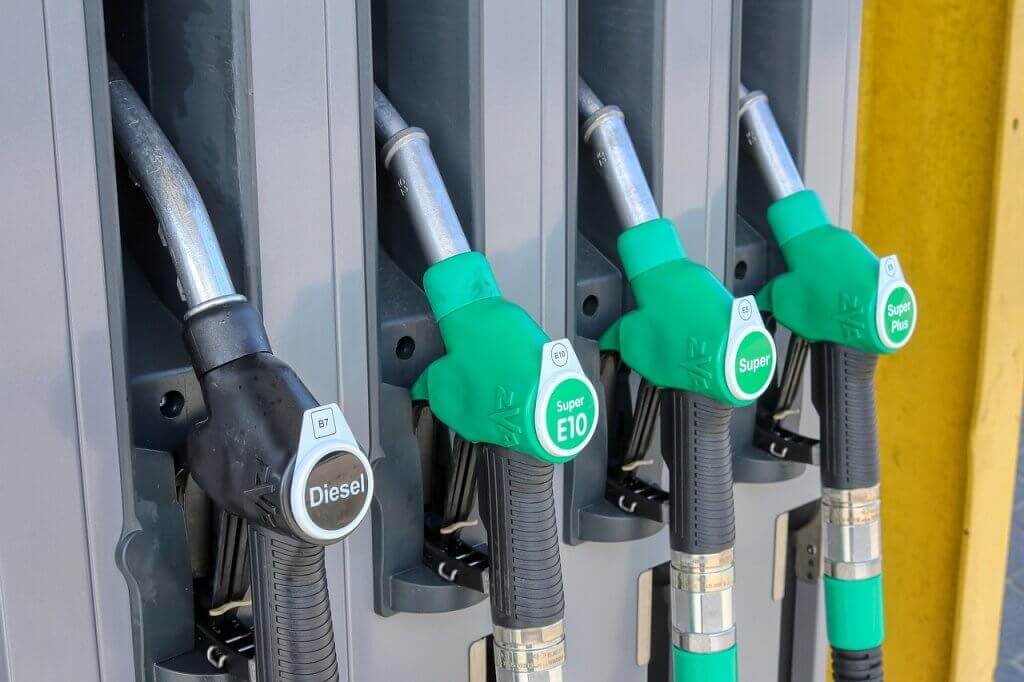 Four fuel pump handles at a gas station: one labeled Diesel, one E10, and two labeled Super Plus, all with green or black handles and hoses attached to a grey fuel dispenser.